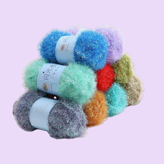 Four balls of fluffy, shimmery yarn in assorted colors including blue, mint green, lavender, red, orange, and gray, stacked together on a white surface with a small decorative rose in the background.