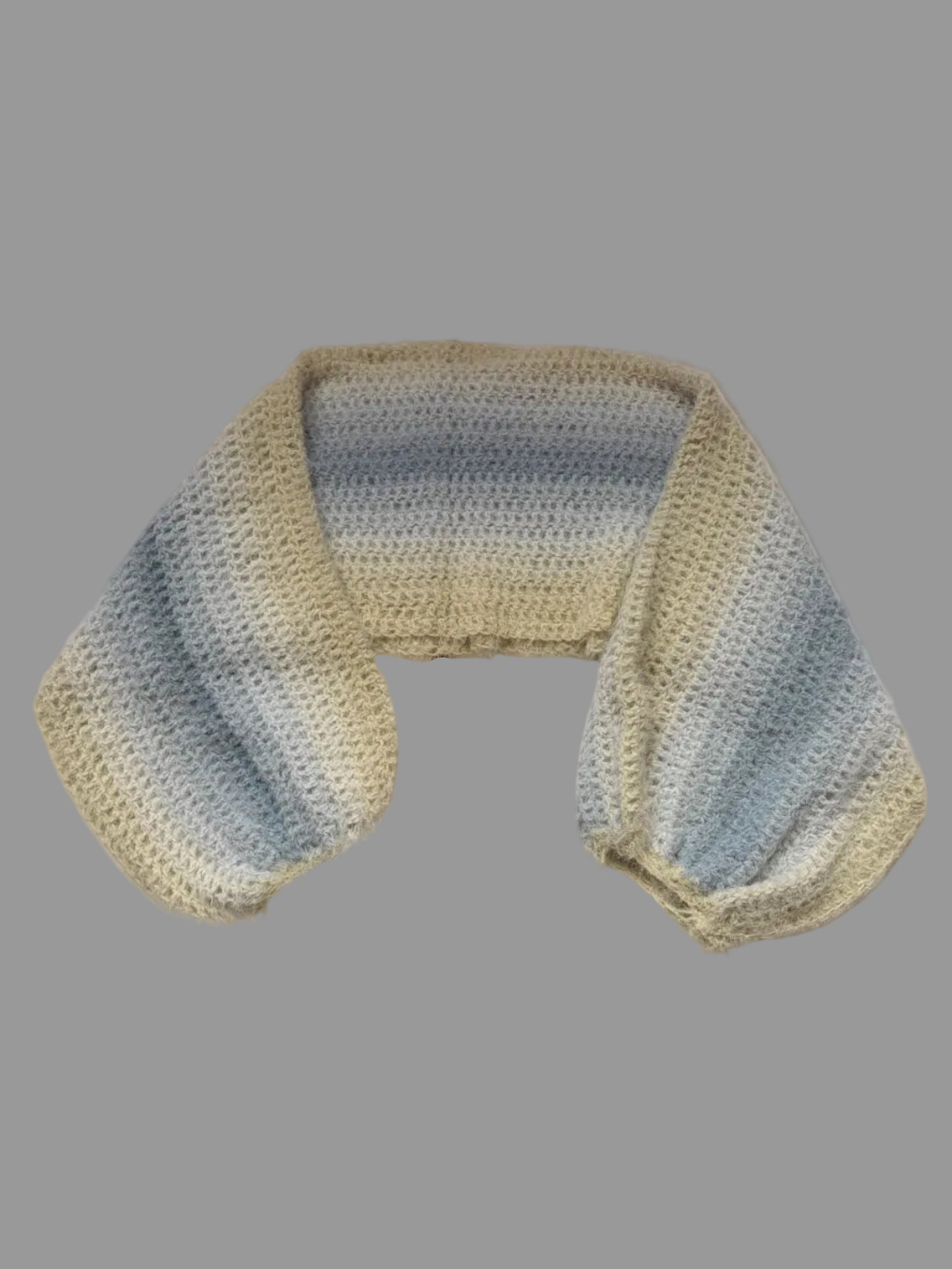 A handmade crochet knit shrug with a striped pattern of light blue, gray, and cream, laid out on a white cabinet.