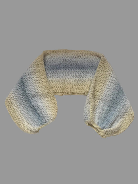 A handmade crochet knit shrug with a striped pattern of light blue, gray, and cream, laid out on a white cabinet.