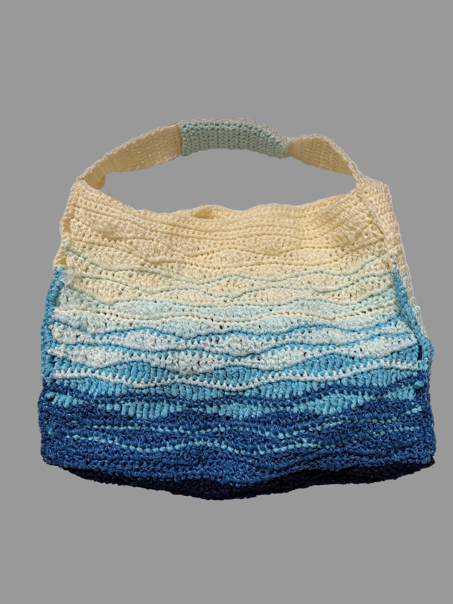 A handmade crochet shoulder tote bag with a wavy horizontal stripe pattern in a gradient of cream, light blue, and dark navy blue, hanging against a white paneled background.