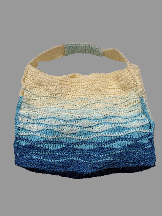 A handmade crochet shoulder tote bag with a wavy horizontal stripe pattern in a gradient of cream, light blue, and dark navy blue, hanging against a white paneled background.